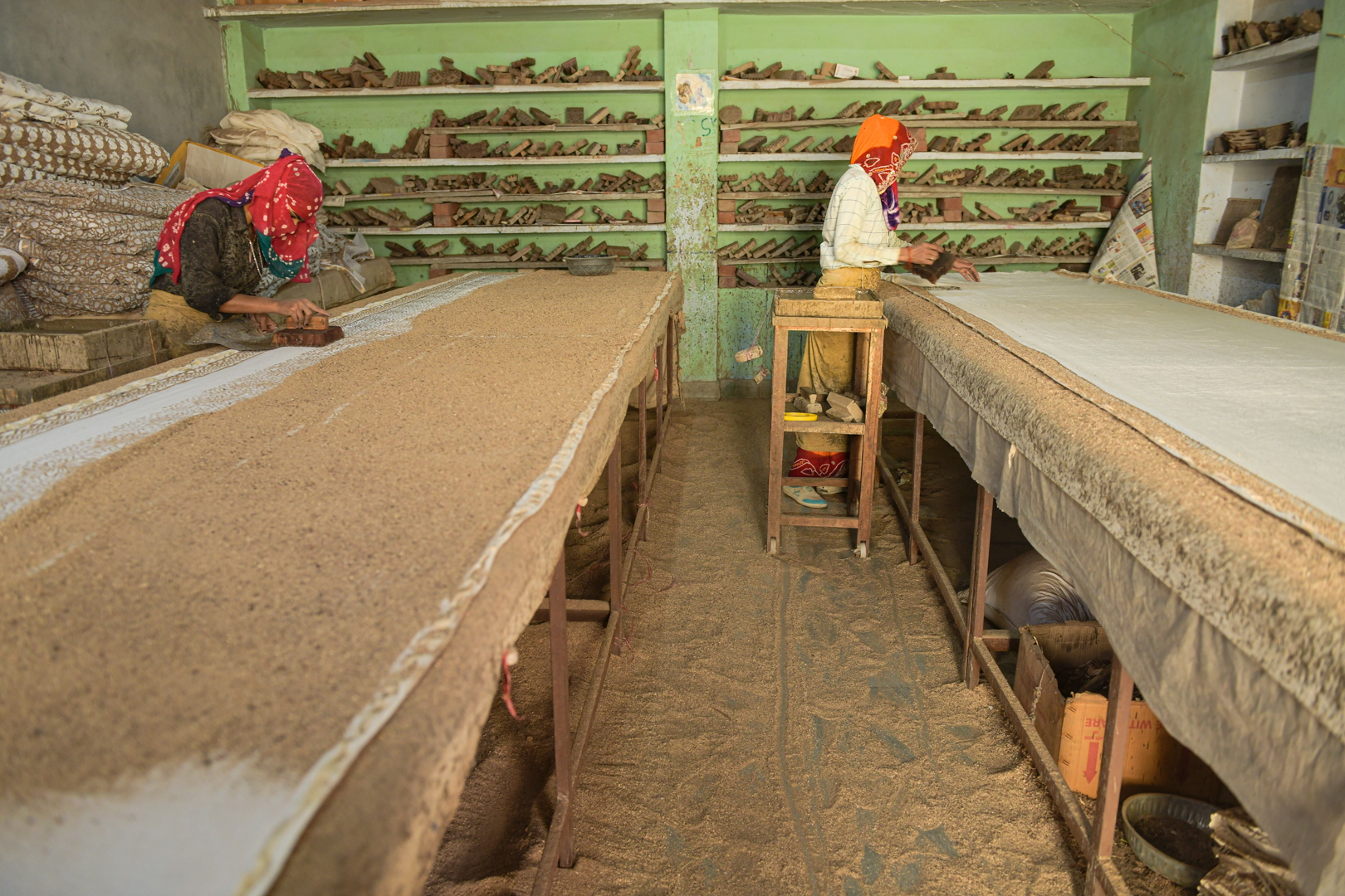 Artisans in a traditional block printing workshop in India meticulously apply patterns to fabric using wooden blocks and mud. Working side by side, they create intricate designs on meters of cloth. Around them, shelves are filled with various blocks that line the walls. Photo Lizane Louw