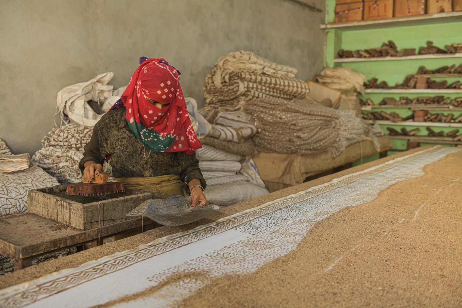 An artisan diligently works on a piece of fabric using a wooden block to apply intricate mud patterns in a traditional block printing workshop in India. Surrounded by stacks of dried textiles and shelves filled with printing blocks, she carefully aligns each block to ensure precision. The detailed process and the rich cultural heritage it represents are evident in her focused efforts and the beautiful designs that emerge. Photo Lizane Louw