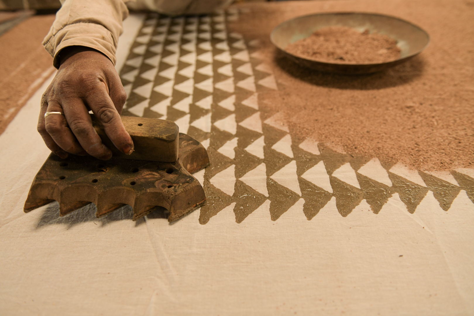 An artisan applies a wooden block to fabric, creating a precise geometric pattern in a traditional block printing workshop in India. The methodical Dabu printing process involves pressing the block, coated with mud, onto the fabric to transfer the intricate design. This close-up image highlights the detailed craftsmanship and skill required in mud block printing. Photo Lizane Louw